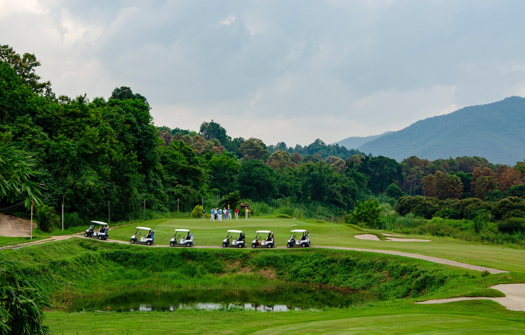 Golfers and carts beside a green surrounded by trees and hills